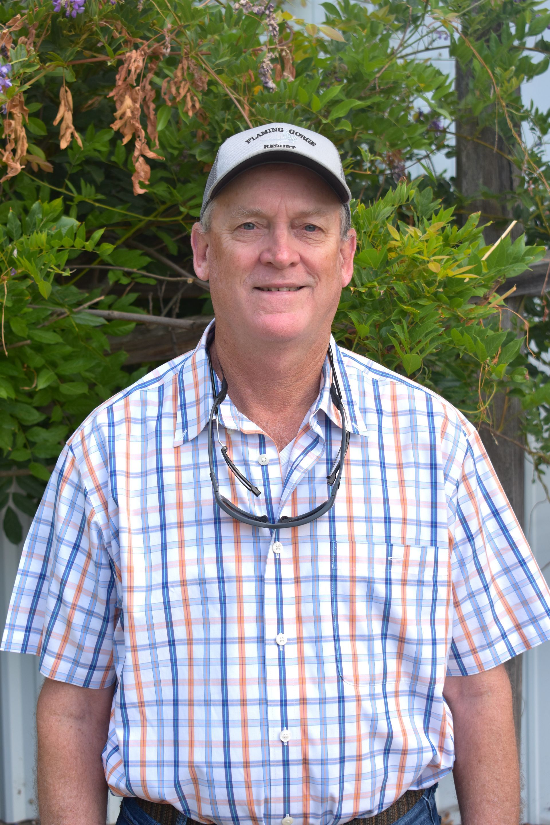 Smiling man in a plaid shirt and cap standing outdoors in front of green foliage.