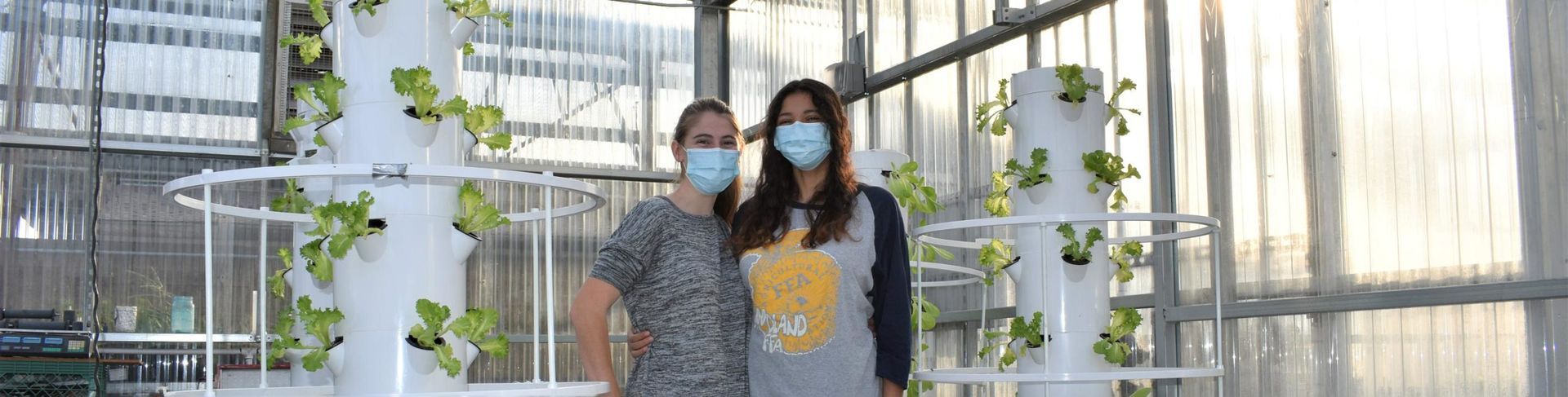 Two people wearing masks pose in a greenhouse with hydroponic towers growing plants.