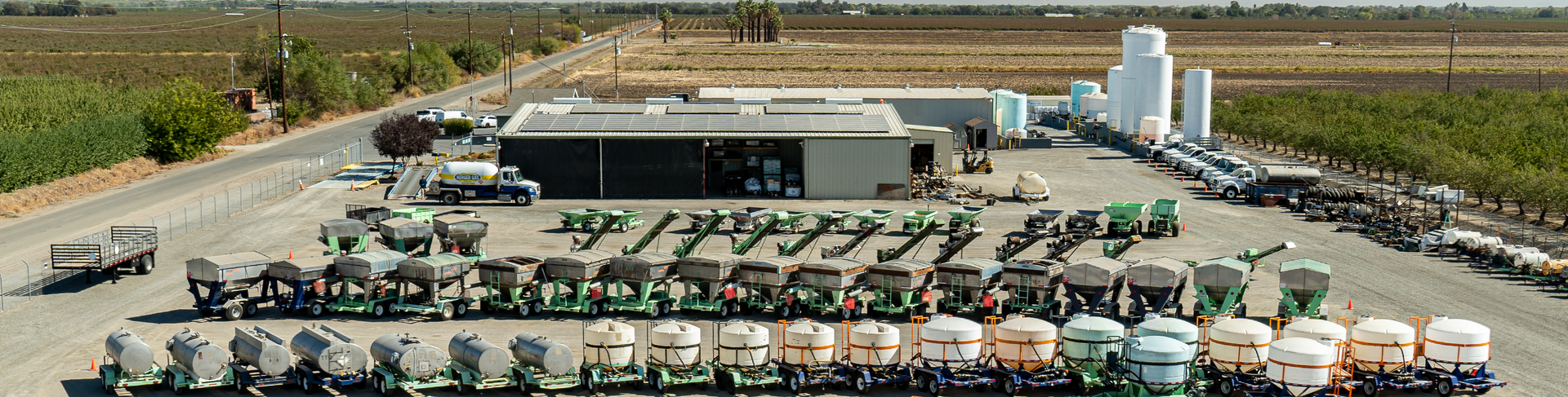 An agricultural facility with many vehicles lined up in a yard. Buildings and silos are in the background.