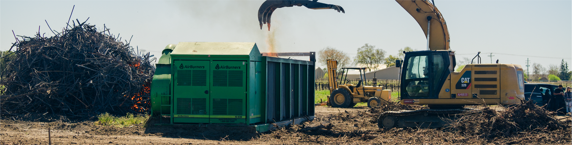 An excavator loads wood chips into a green machine, next to a pile of brush, outdoors.