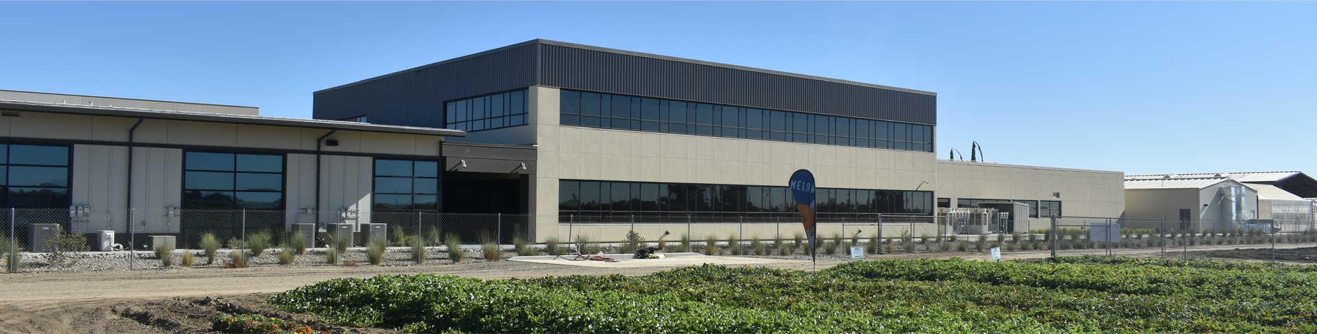 Modern industrial building with multiple stories and windows against a blue sky, fronted by a cultivated field.