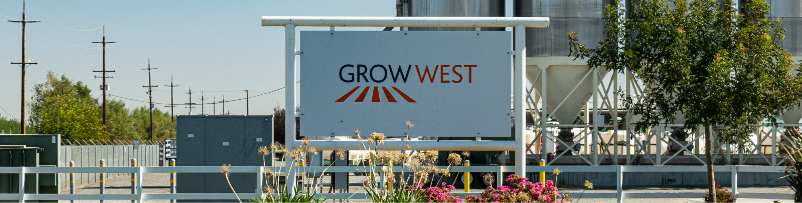 Sign for Grow West. The sign is white with the company logo. Buildings, trees and power lines are visible in the background.
