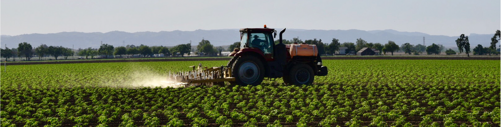 A tractor sprays crops in a green field with a distant mountain range in the background.