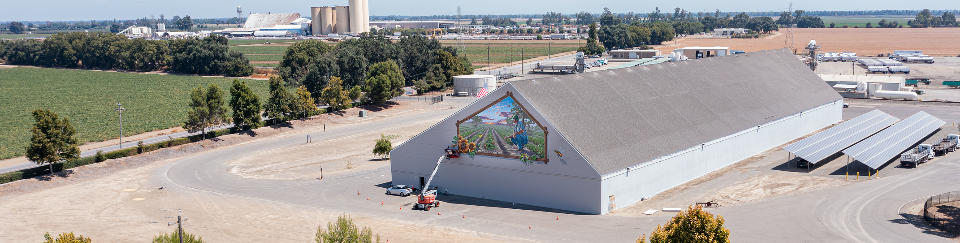 Aerial view of a large building with a mural, farmland, solar panels, and silos in the background.