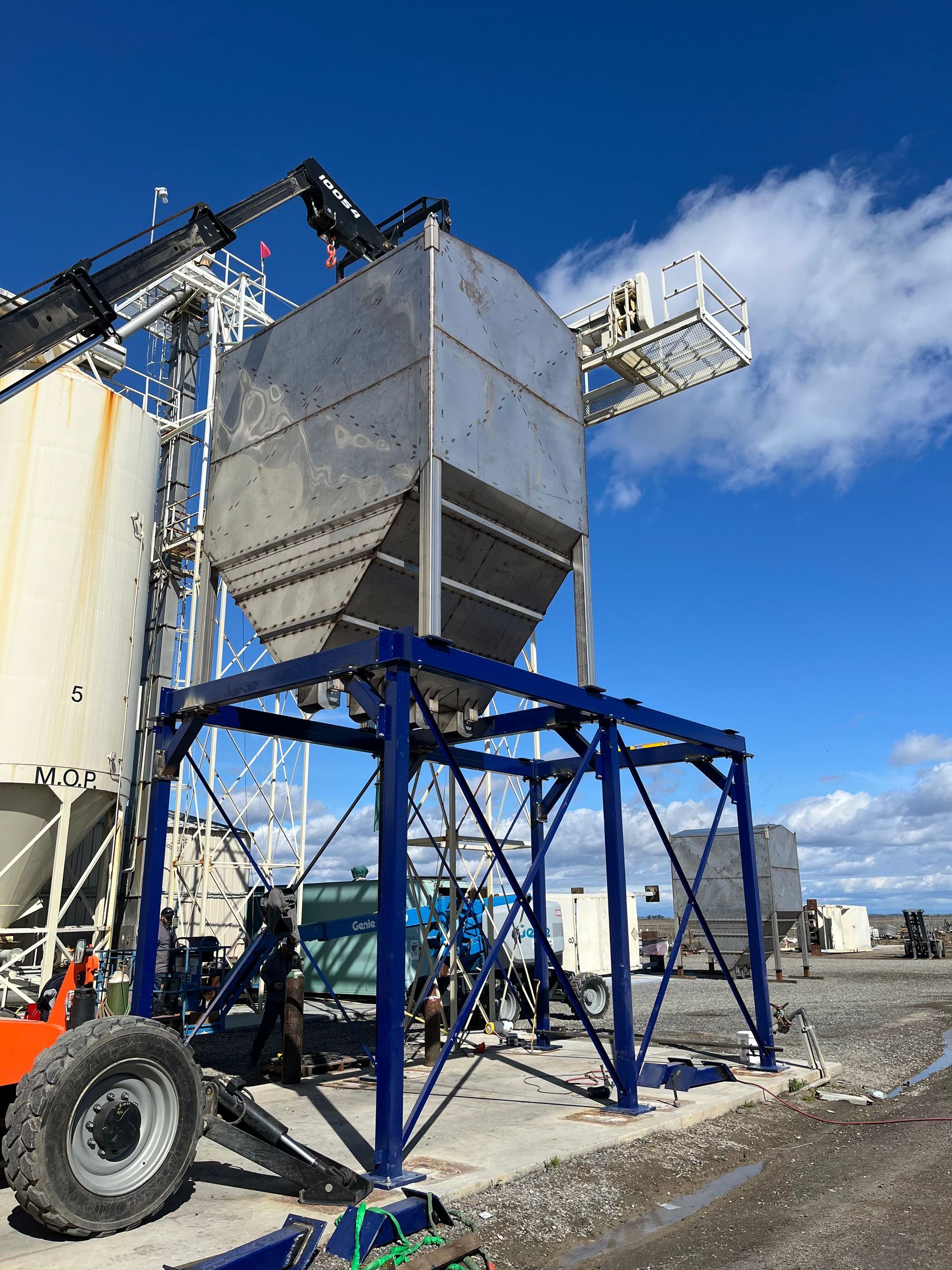 Stainless steel industrial hopper on a blue steel support frame under a bright blue sky.