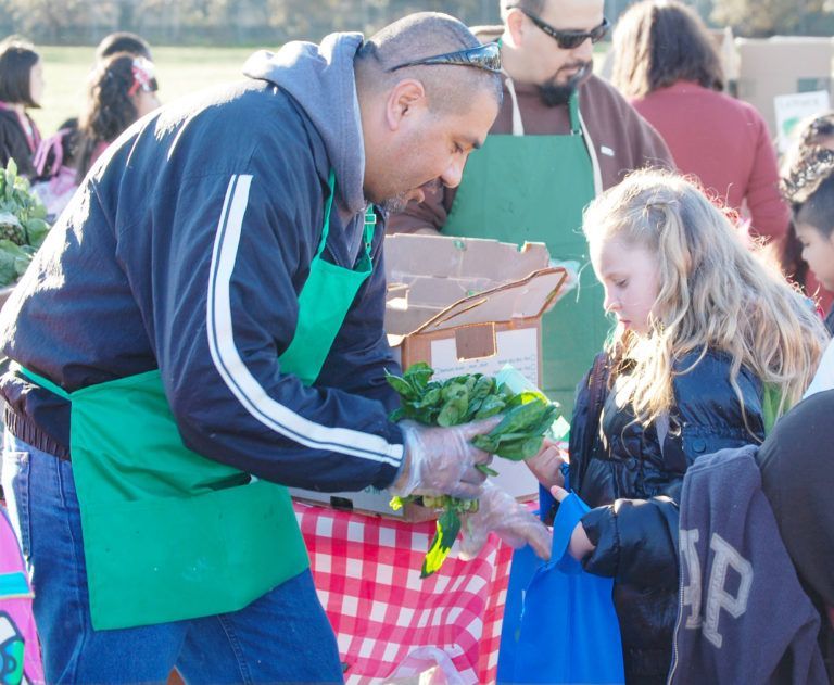 Man in apron handing produce to child at outdoor market.