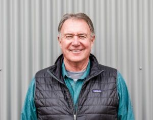 Man wearing a black vest and turquoise shirt smiles in front of a corrugated metal wall.