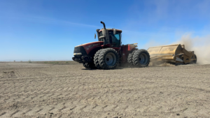 Red tractor pulling a scraper across a dry, dusty field under a blue sky.