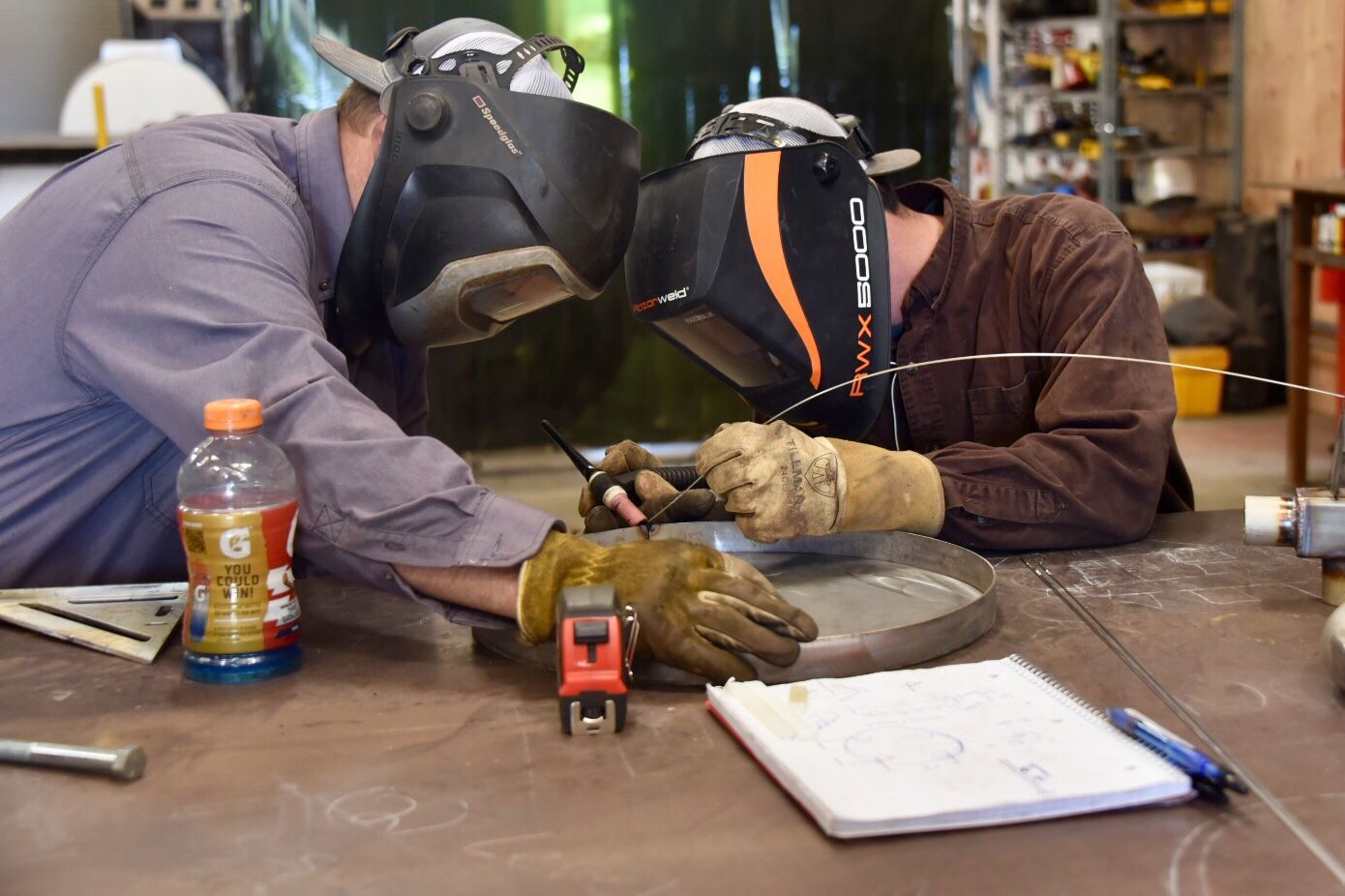 Two welders in protective gear work on metal at a workbench indoors.