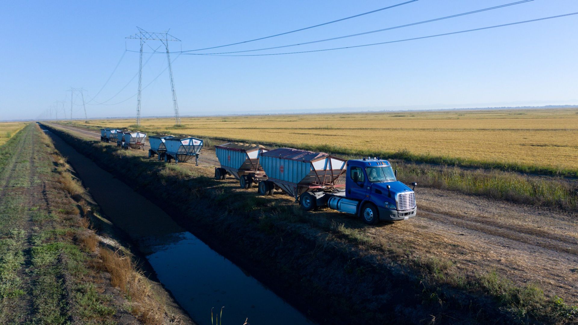 Blue truck pulling trailers along a dirt path beside a canal and harvested fields under power lines.