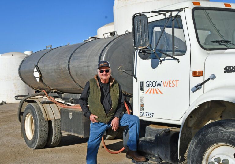 Man stands next to Grow West tanker truck. He wears a cap, vest, and jeans. Bright, sunny day.