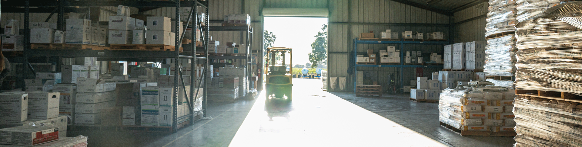 A warehouse interior with boxes on shelves and a forklift in the aisle, bright light at the open door.