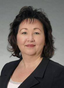 Woman with dark hair in a black blazer smiling, against a gray gradient background.