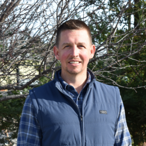 Man wearing a blue vest and plaid shirt smiles in front of a tree.