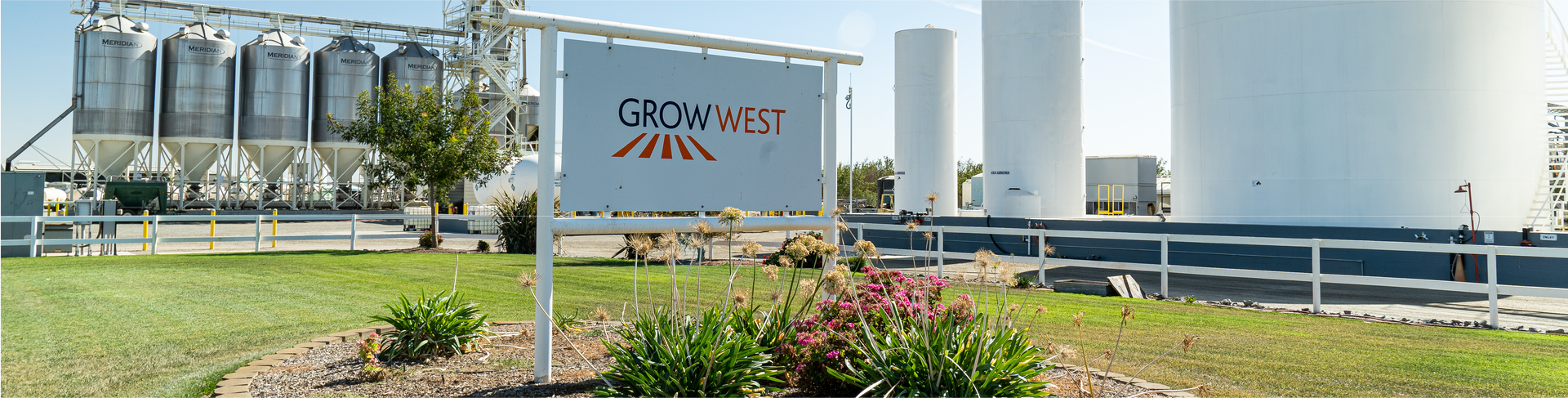 Sign for Grow West agricultural facility with silos and tanks in background.