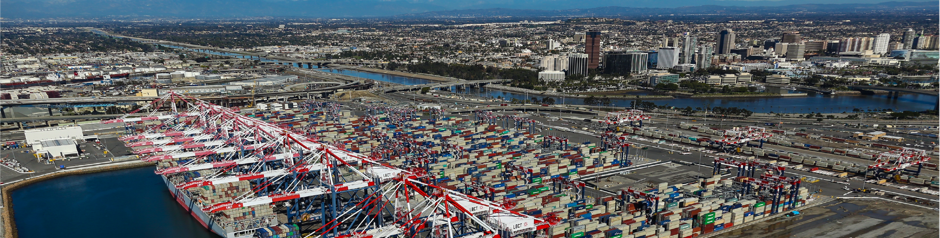 Aerial view of a busy port with container ships, cranes, and surrounding city buildings under a blue sky.