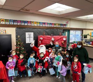 Santa Claus with children at a holiday event, all wearing masks, receiving gifts in a classroom.