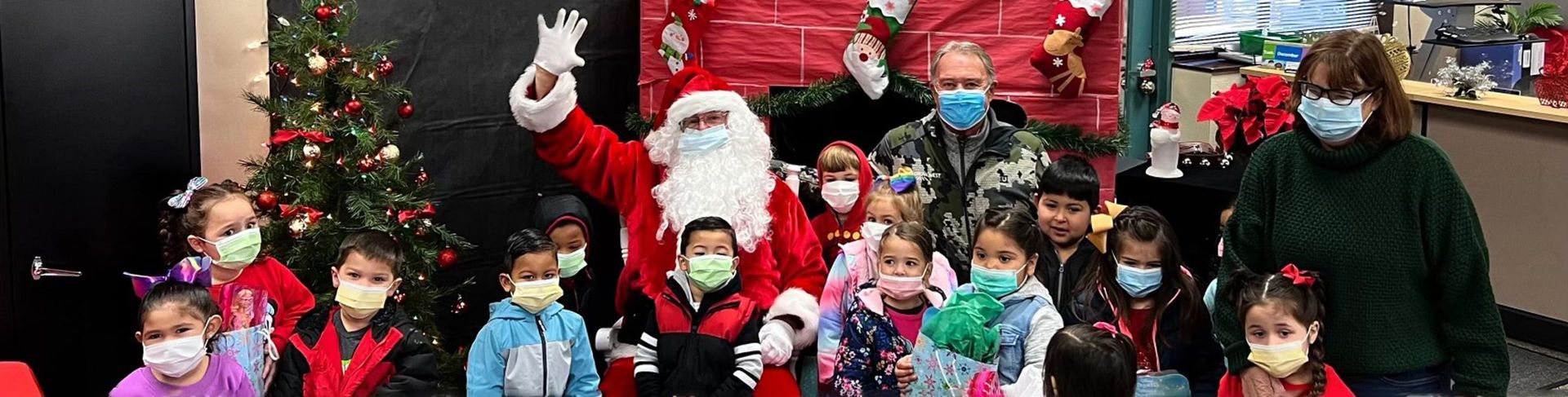 Santa Claus with children and adults wearing face masks pose together near a Christmas tree.
