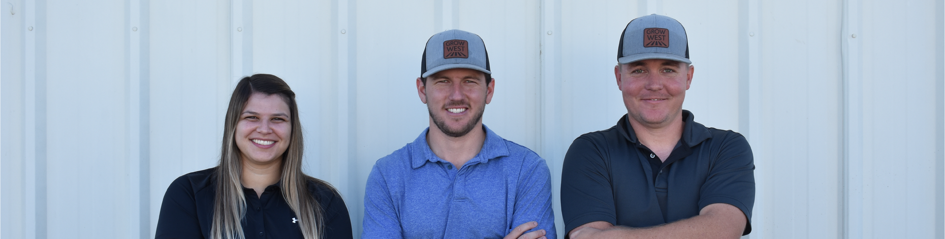 Three people smiling in front of a white wall; two men wearing hats, and the other person is a woman.