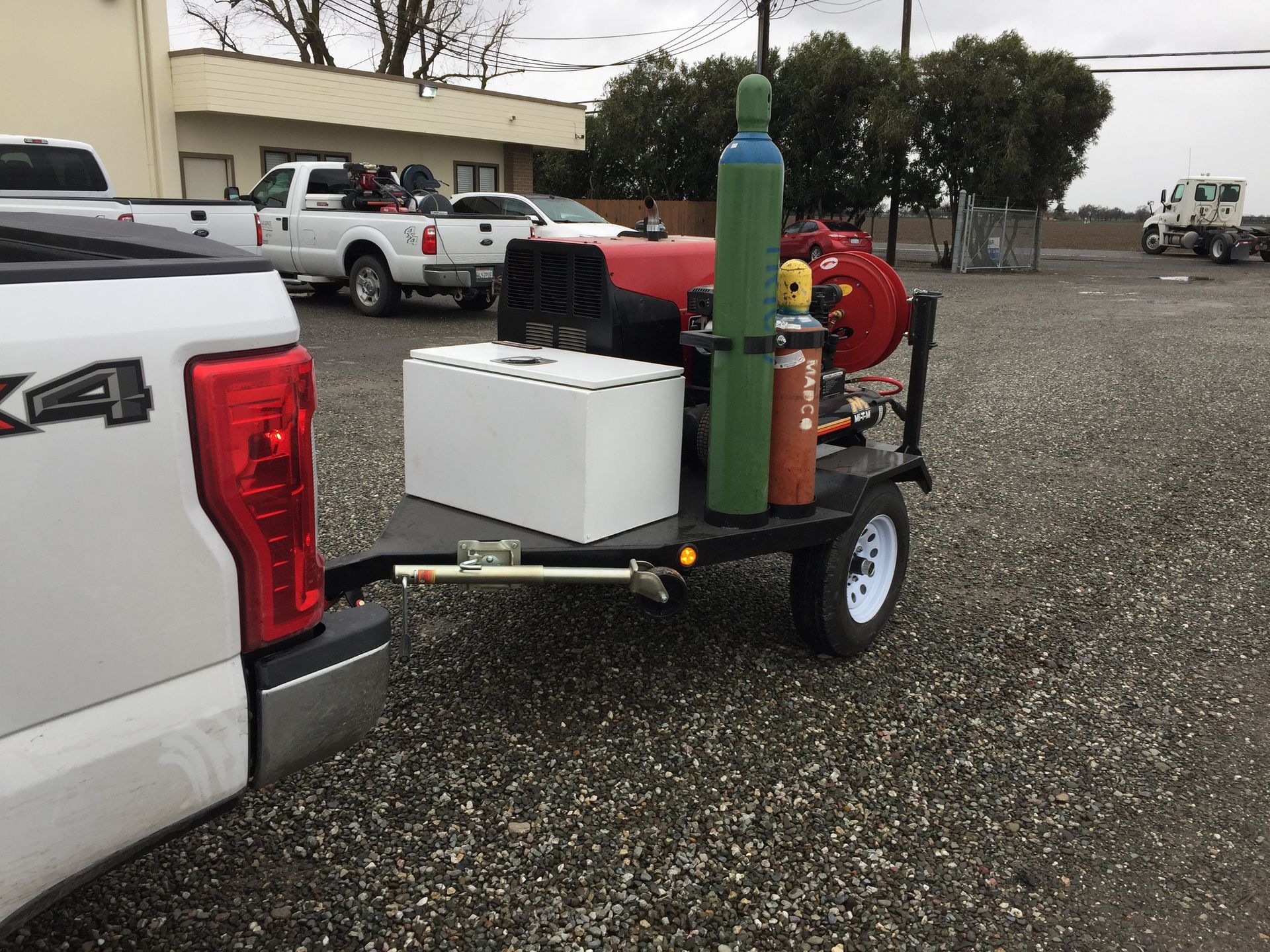 Trailer with a white cooler and gas tanks parked on gravel beside a white pickup truck.