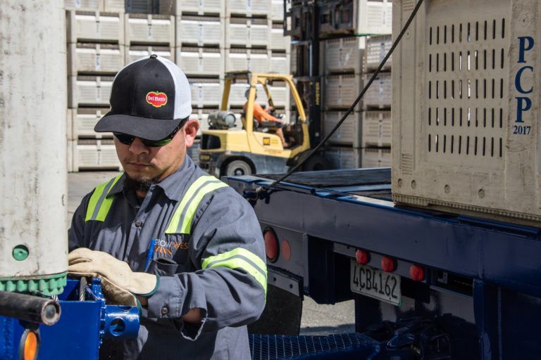 Man in work clothes loads crate onto truck near a forklift and stacked crates.