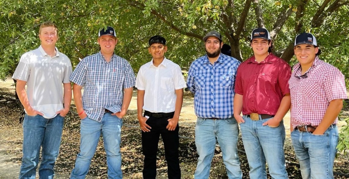 Six young men in casual attire posing outdoors. They are standing under trees, looking at the camera.