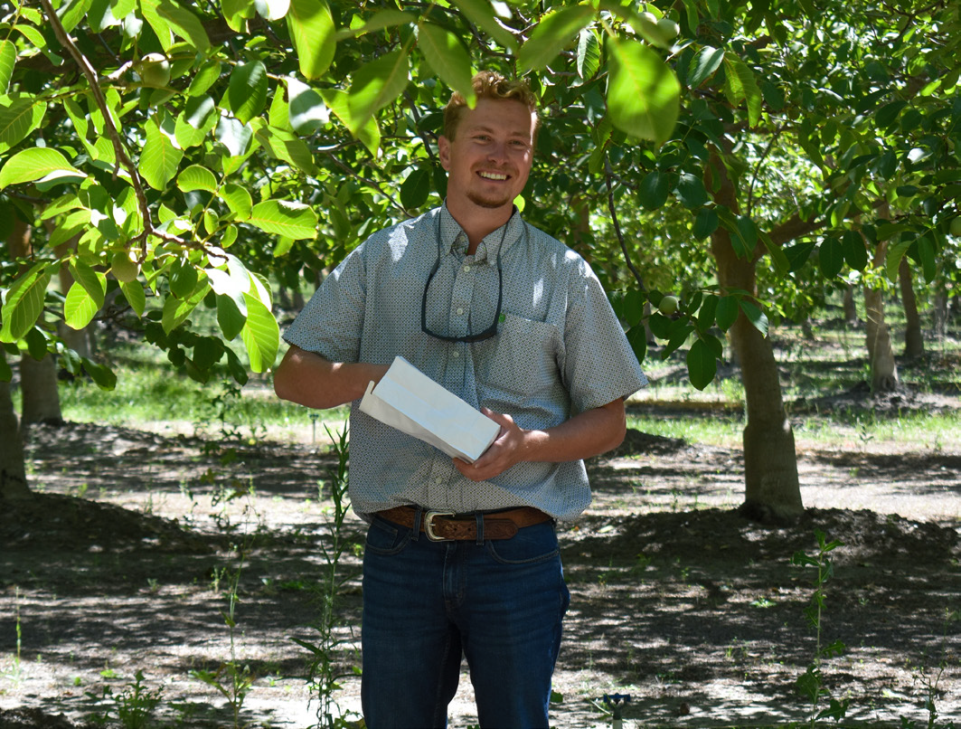 Man holding papers, standing in orchard, smiling.