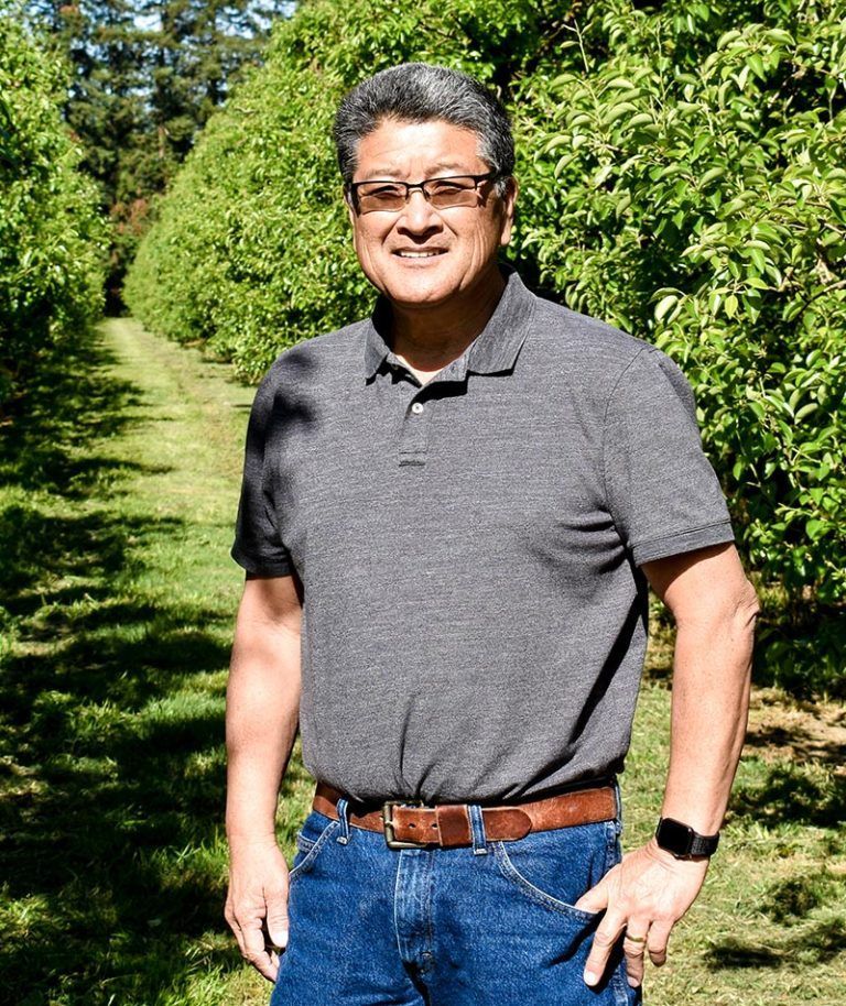 Man in gray shirt and jeans standing in an orchard, smiling.