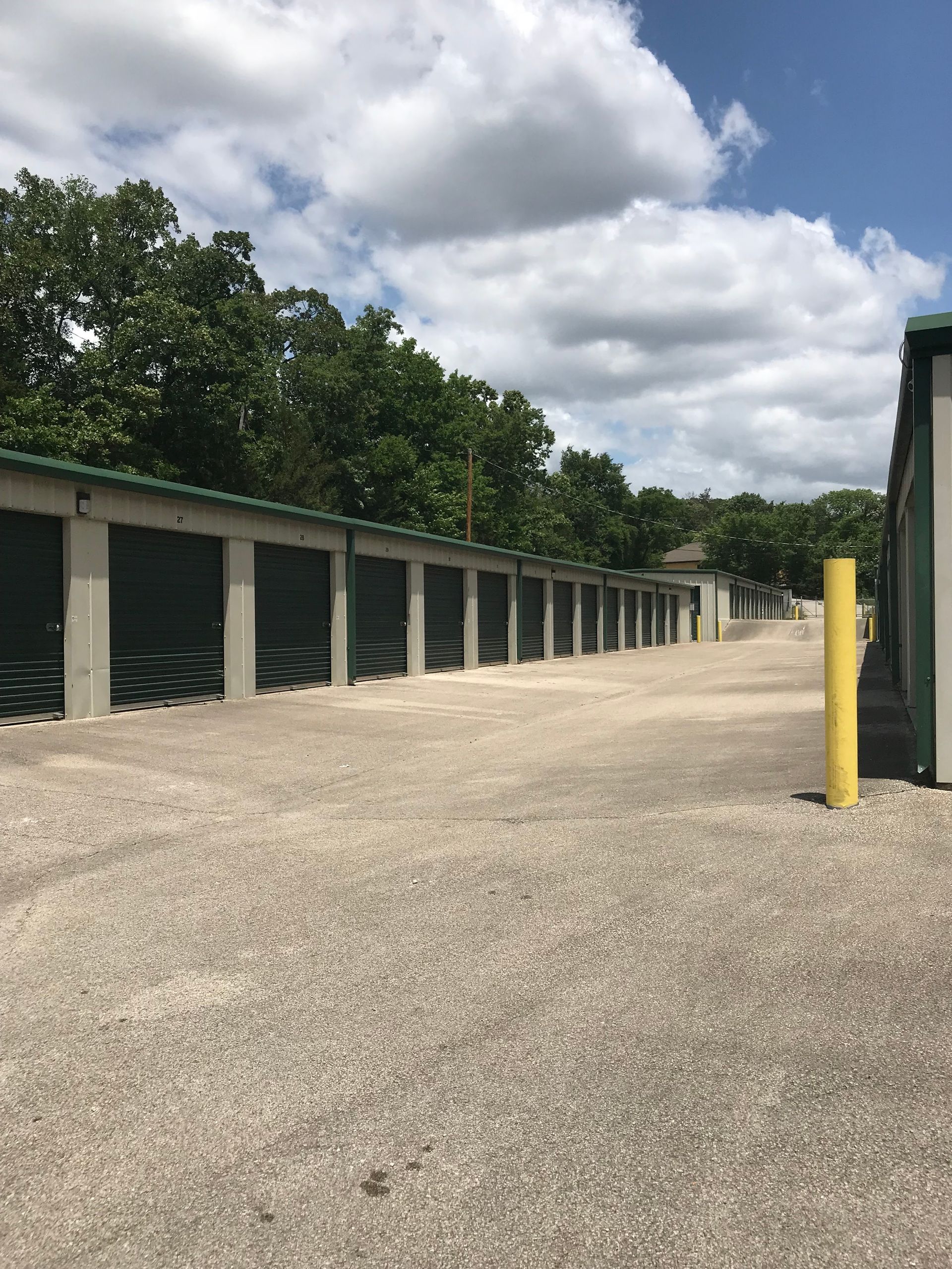 Outdoor storage units with green doors beside a gravel lot and yellow post under cloudy sky