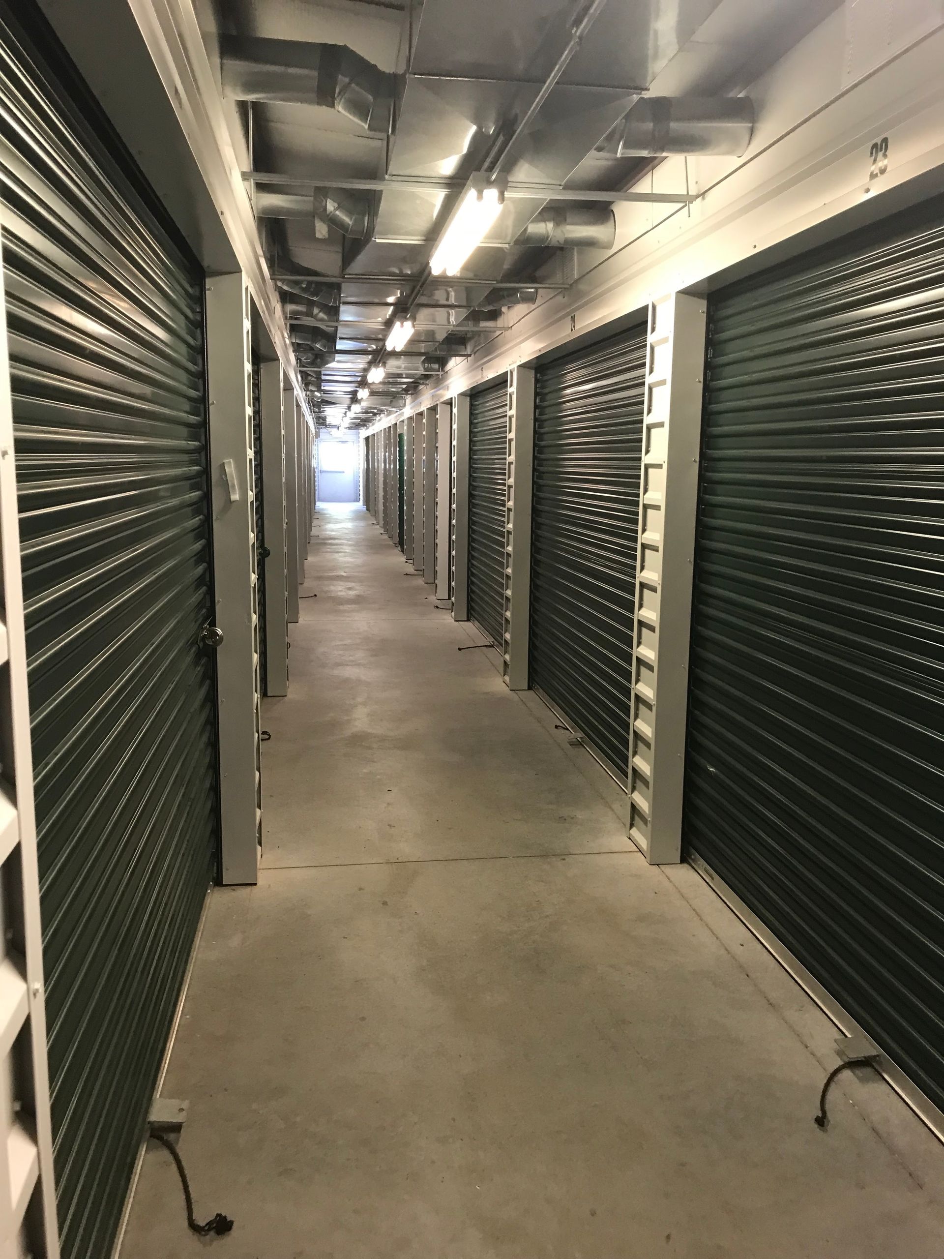 Long storage corridor with rows of dark metal shutters and overhead pipes, lit by fluorescent lights.