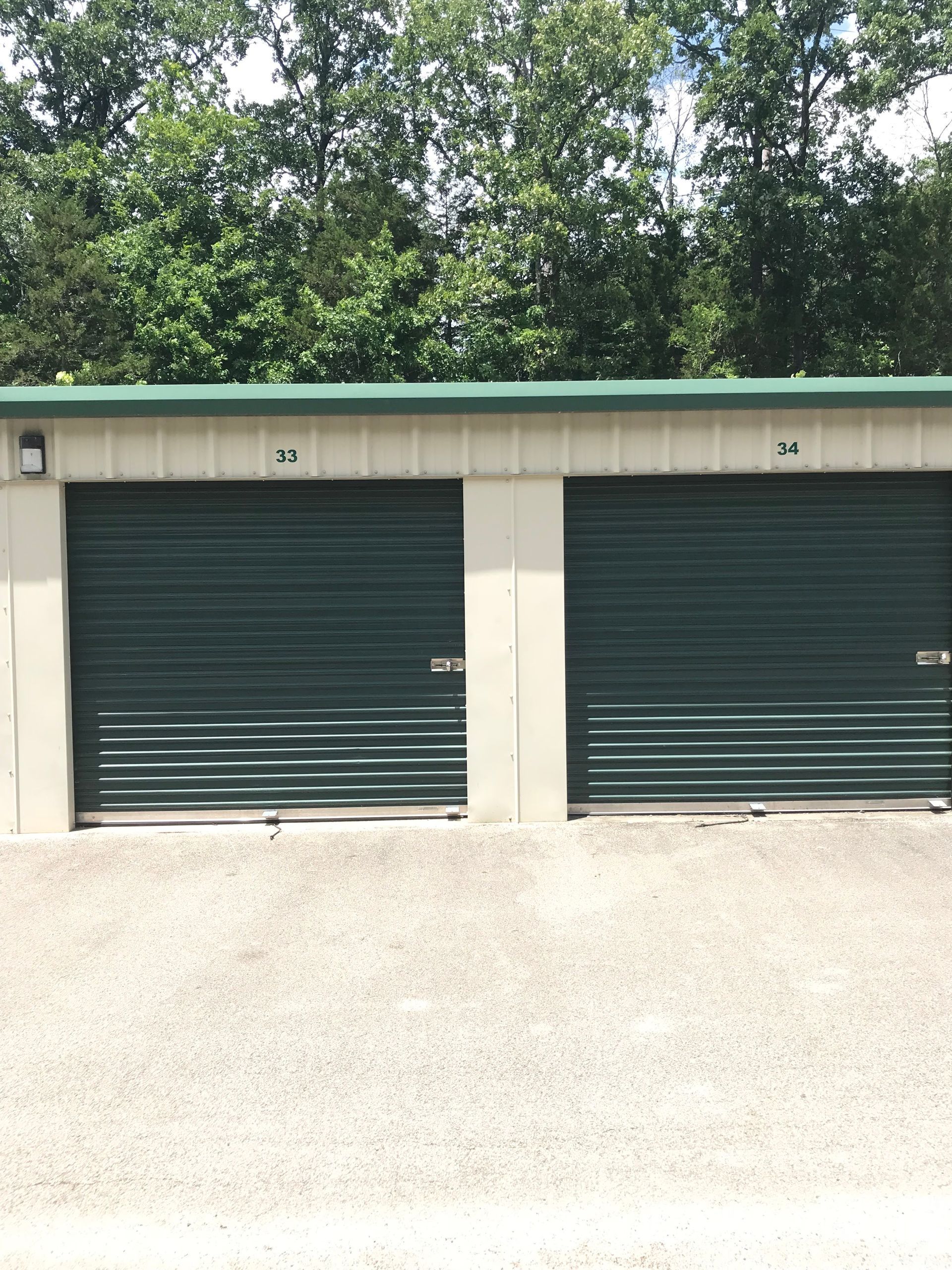 Two green storage unit doors in a row beneath a green roof, with gravel foreground and trees behind.