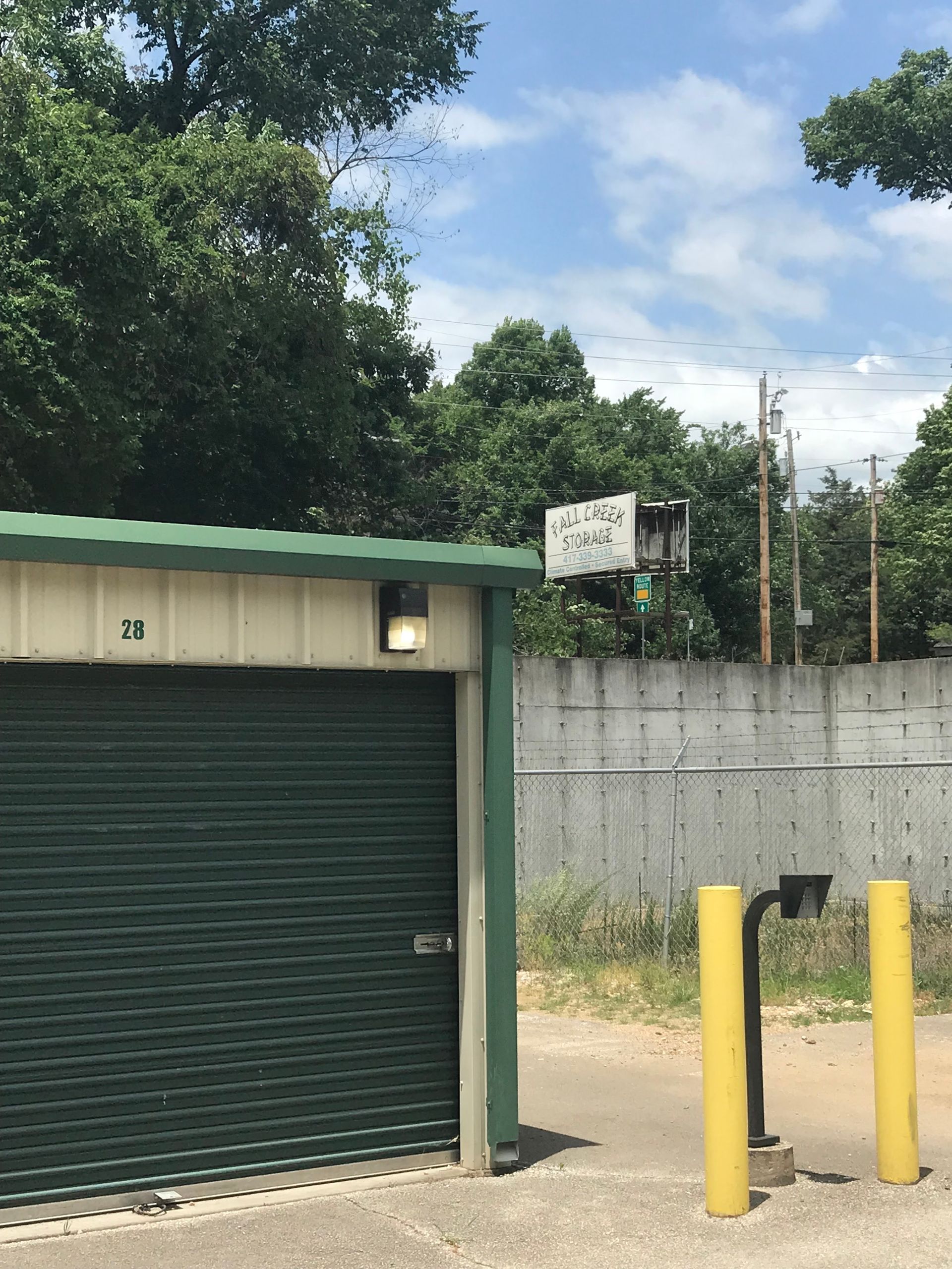 Green roll-up garage door beside concrete wall and yellow bollards under a partly cloudy sky.