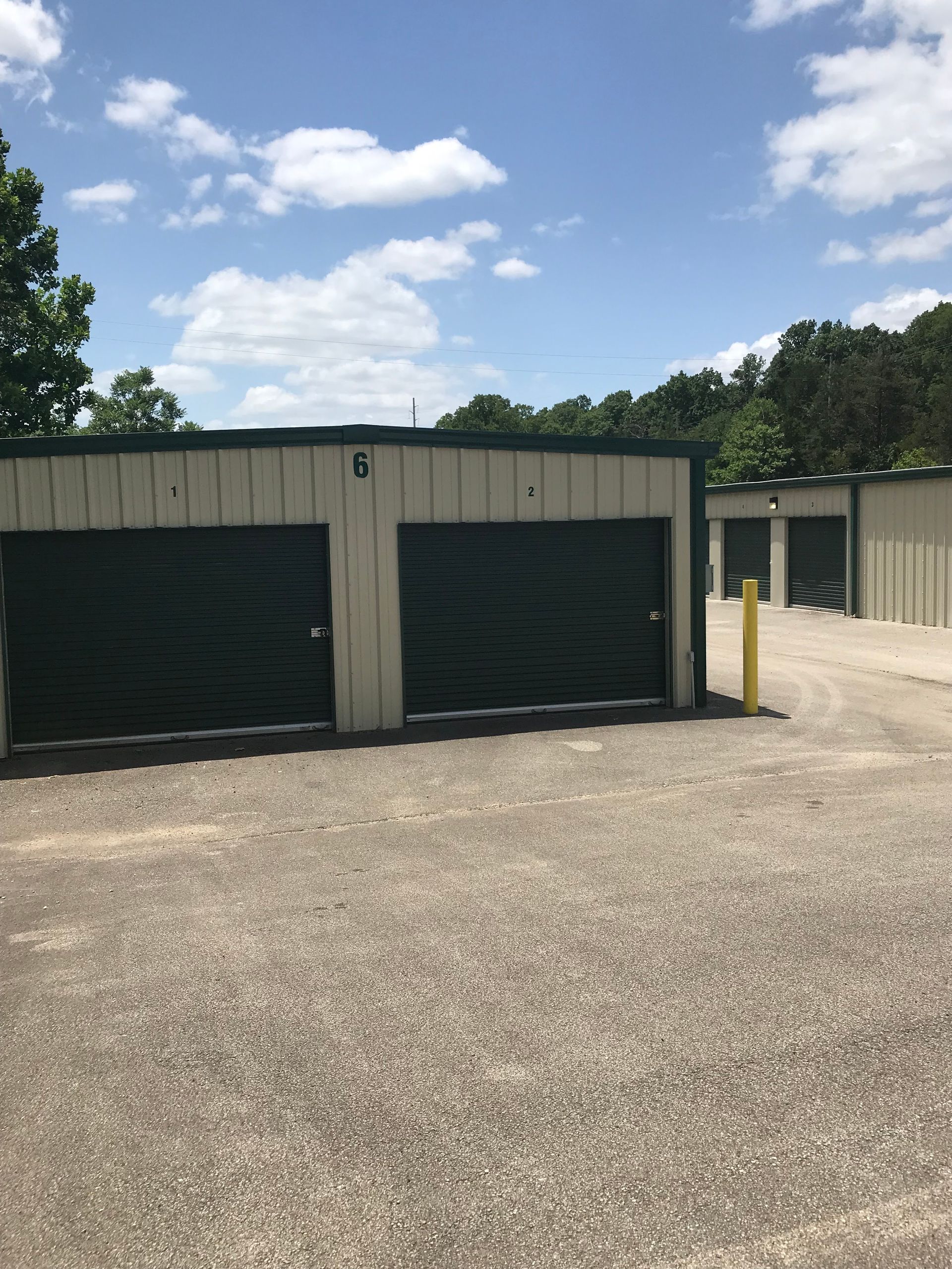 Two beige storage units with dark green doors on a gravel lot under a blue sky