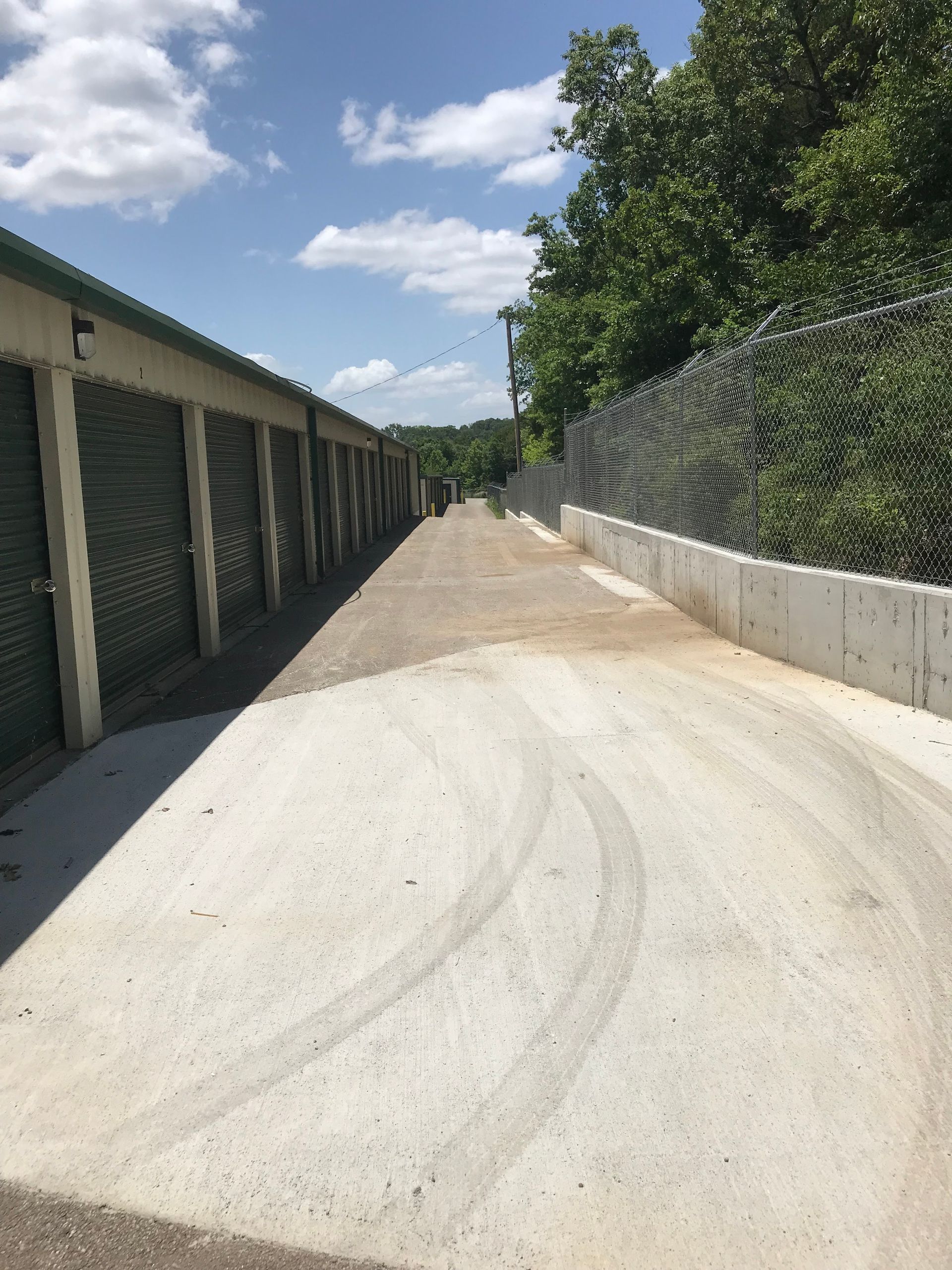 Sunny storage-unit alley with concrete driveway, chain-link fence, and trees on the right