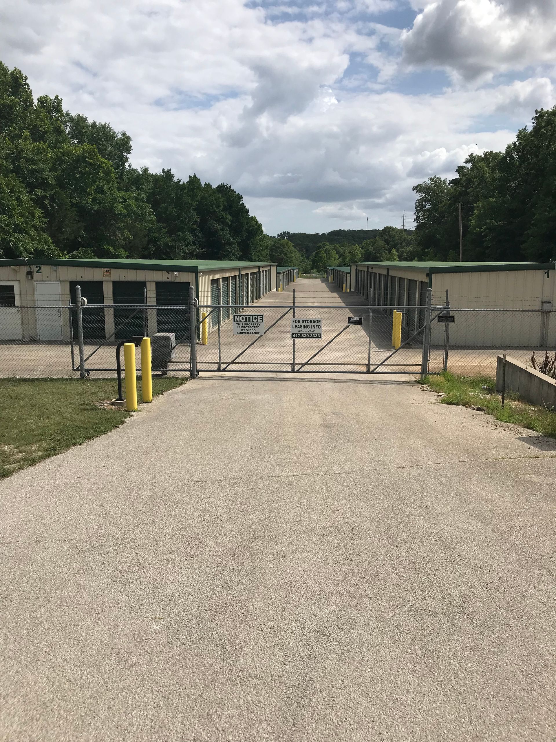 Gravel driveway leading to a closed chain-link gate between two concrete buildings with trees beyond