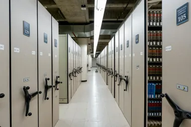 Rows of beige archive cabinets in a storage room, with hand cranks and shelves of files visible.