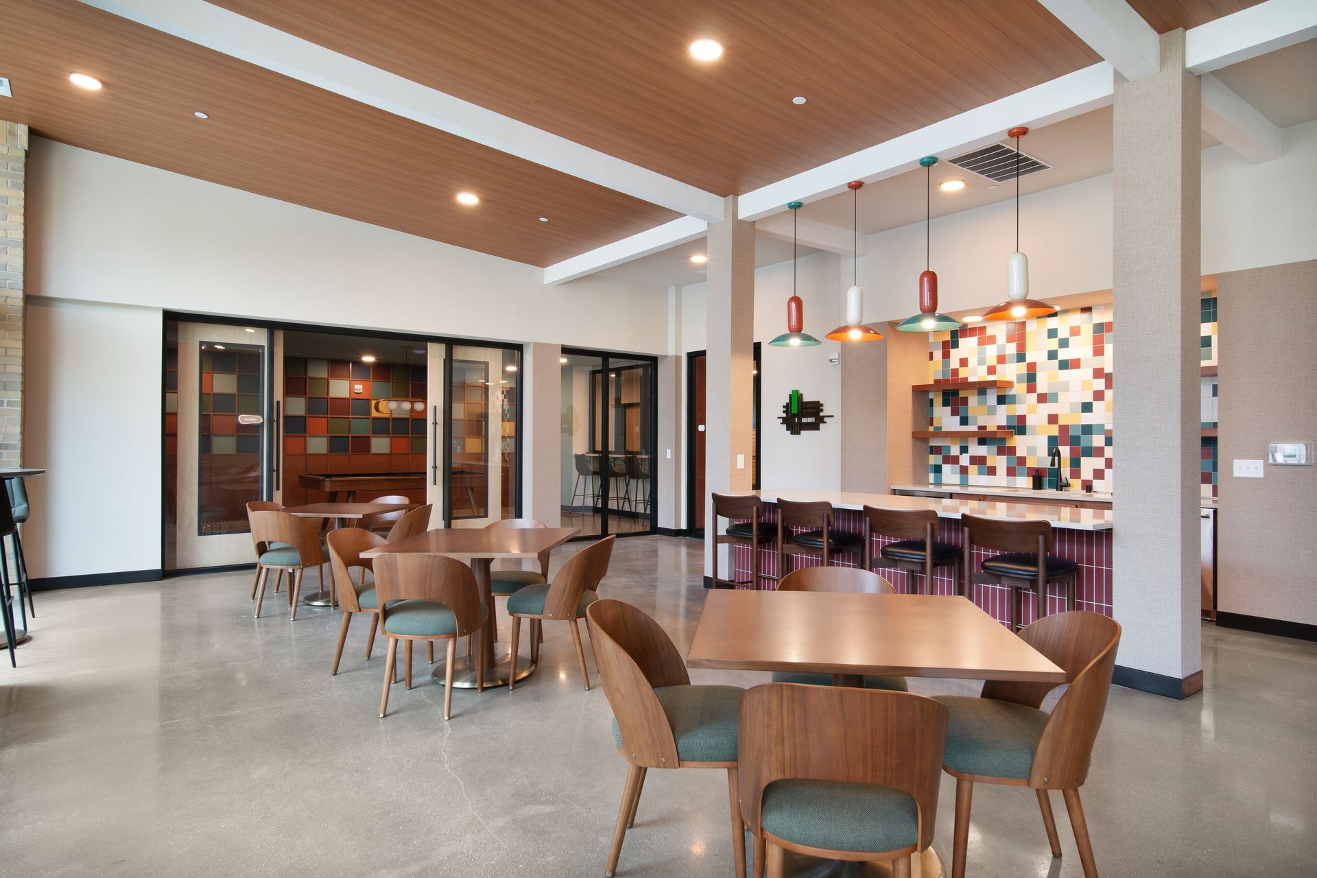 Modern lounge with wooden ceiling, round tables, chairs, colorful wall art, and glass doors.