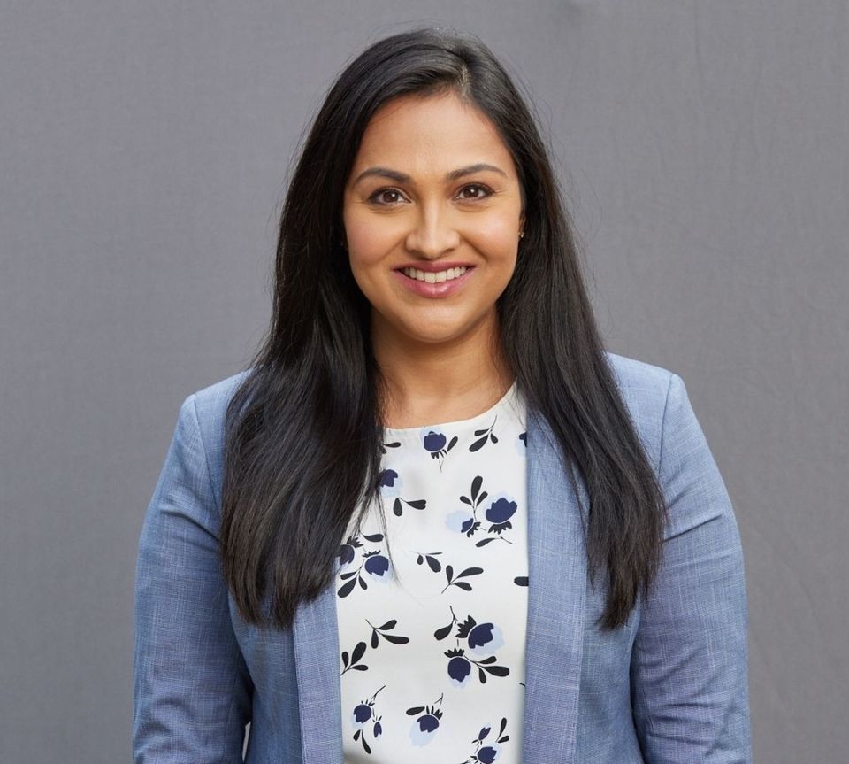 Woman with dark hair smiles, wearing a blue blazer and floral print shirt, against a gray backdrop.