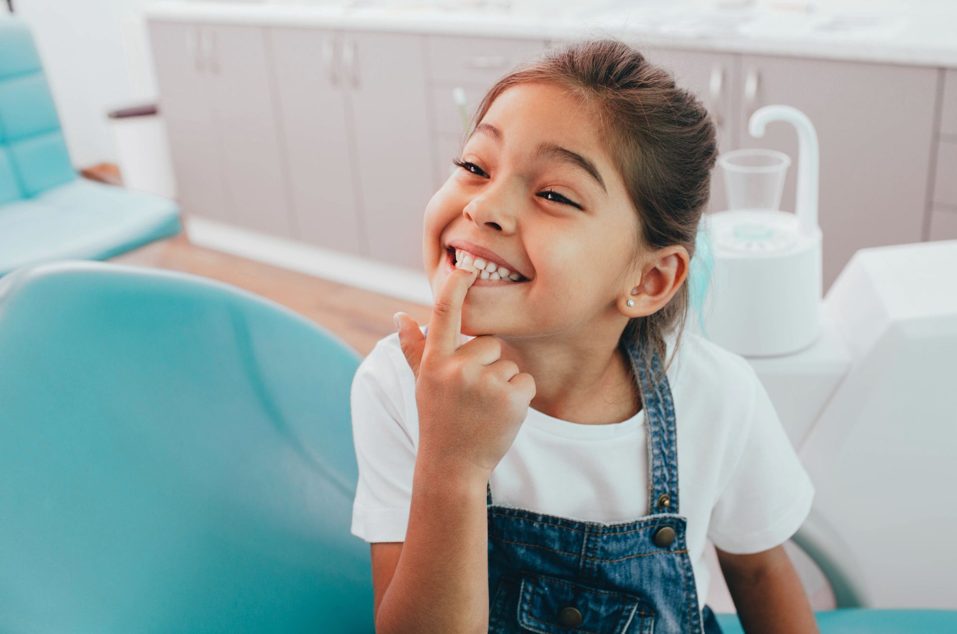 A little girl is sitting in a dental chair and pointing at her teeth.