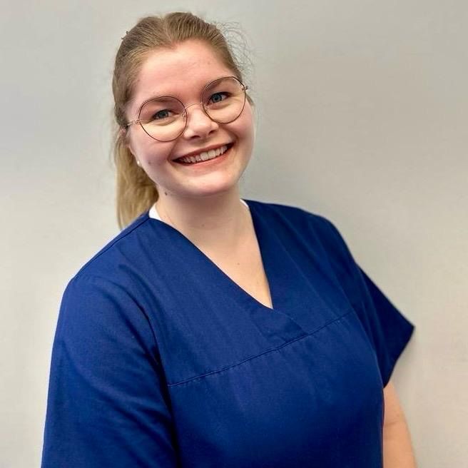 Une personne souriante portant des lunettes et une blouse médicale blanche à bordure orange se détache sur un fond clair uni.