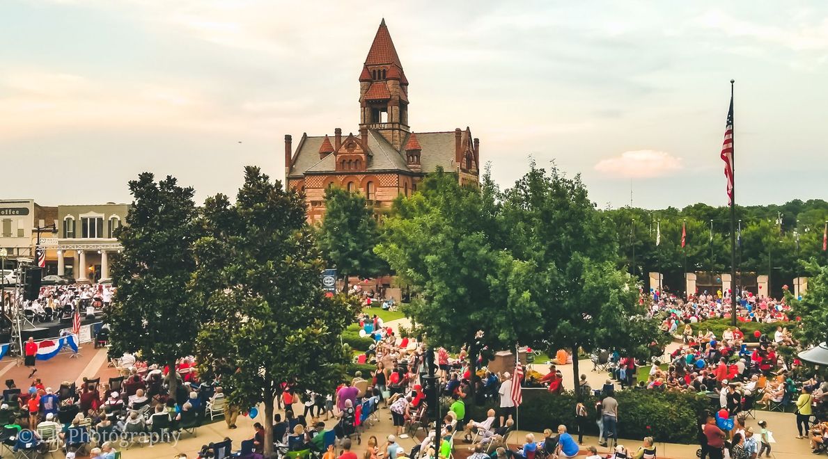 Crowd gathered in a park before a tall historic church or tower, with trees and flags in the background