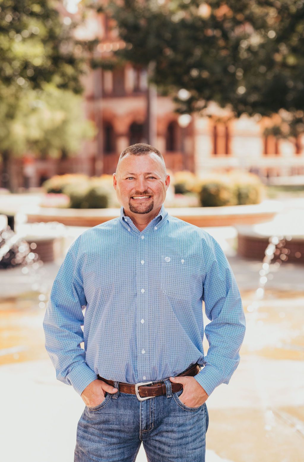 Man in blue patterned shirt standing outdoors in front of a fountain, smiling
