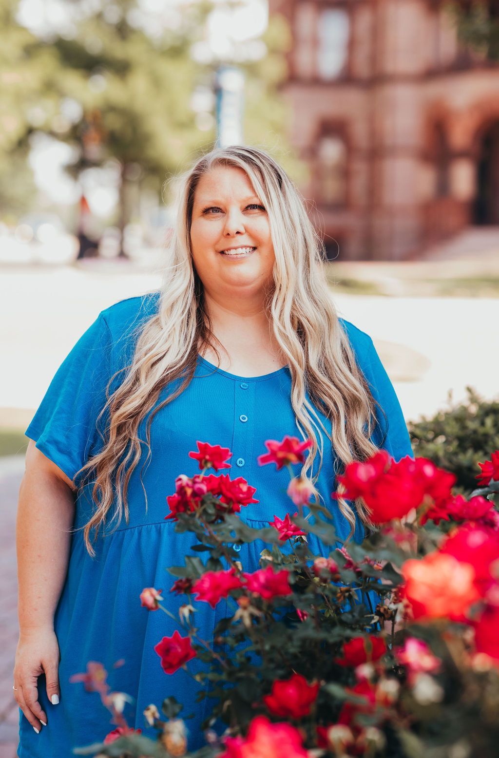 Smiling woman in a blue dress standing outdoors beside bright red flowers