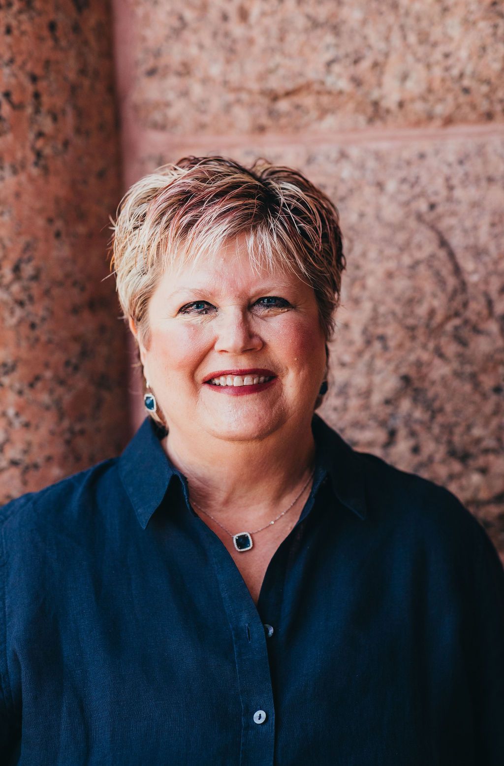 Smiling woman in a dark blue shirt standing against a pink stone wall