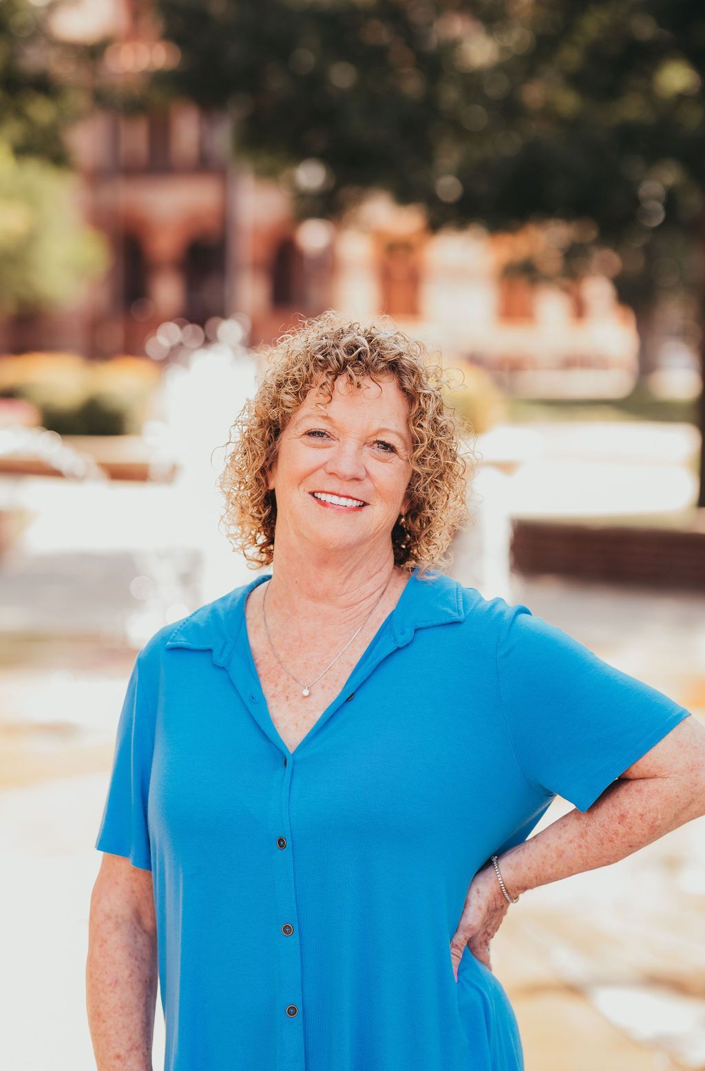 Smiling person in a bright blue shirt standing outdoors with a blurred fountain and building behind them