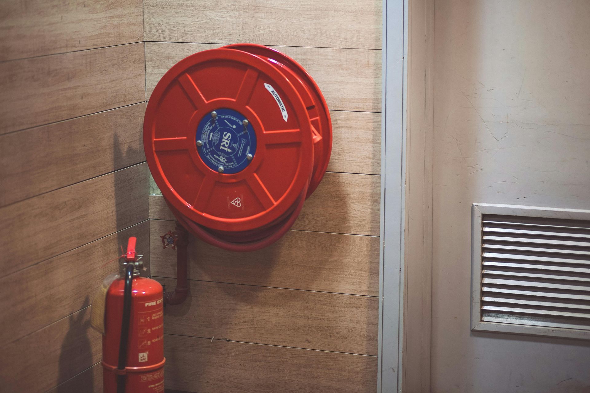 Red fire hose reel and extinguisher mounted on a wooden wall beside a vented door panel