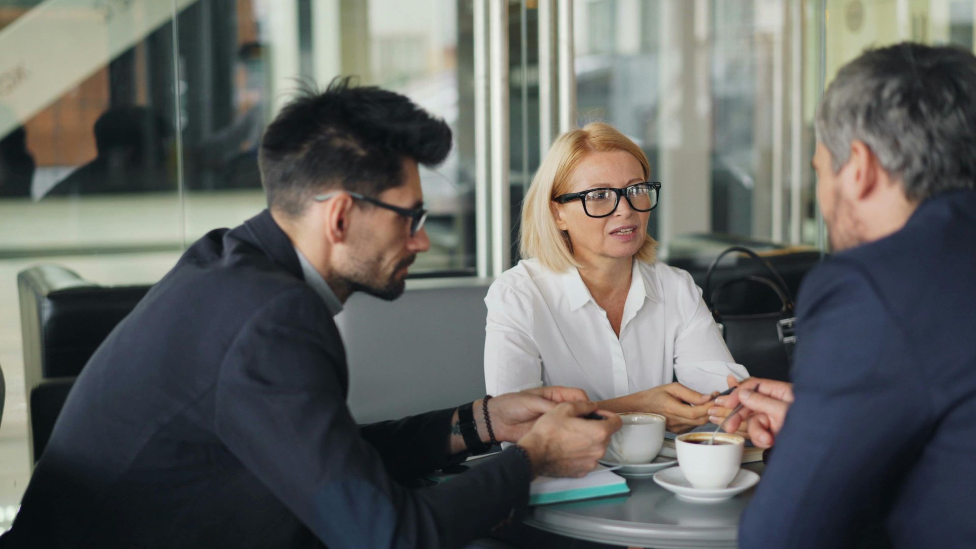 Three professionals in a meeting at a conference table, discussing documents and coffee cups.