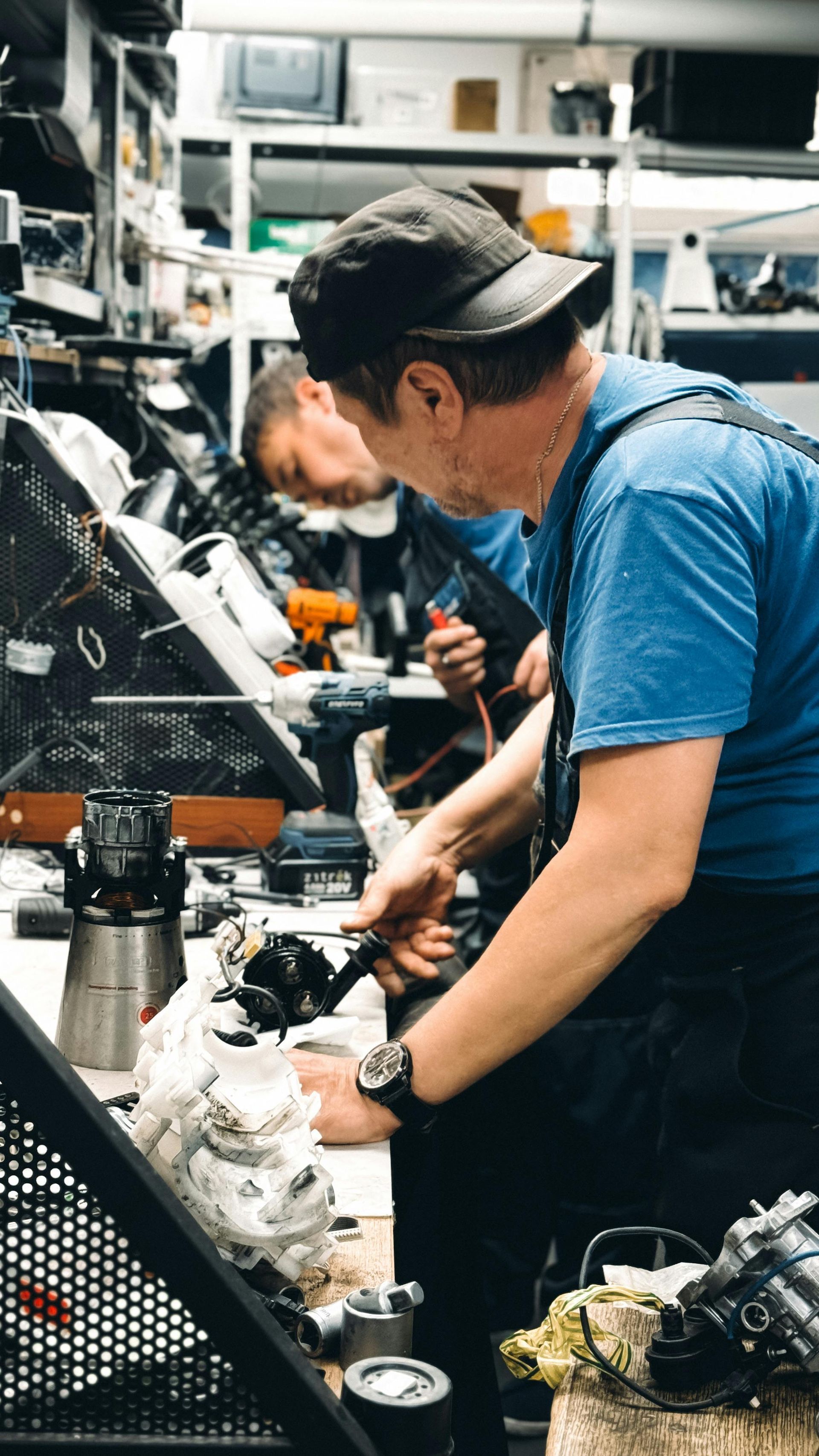 Two people working on machinery in a cluttered workshop, one in a blue shirt and black cap