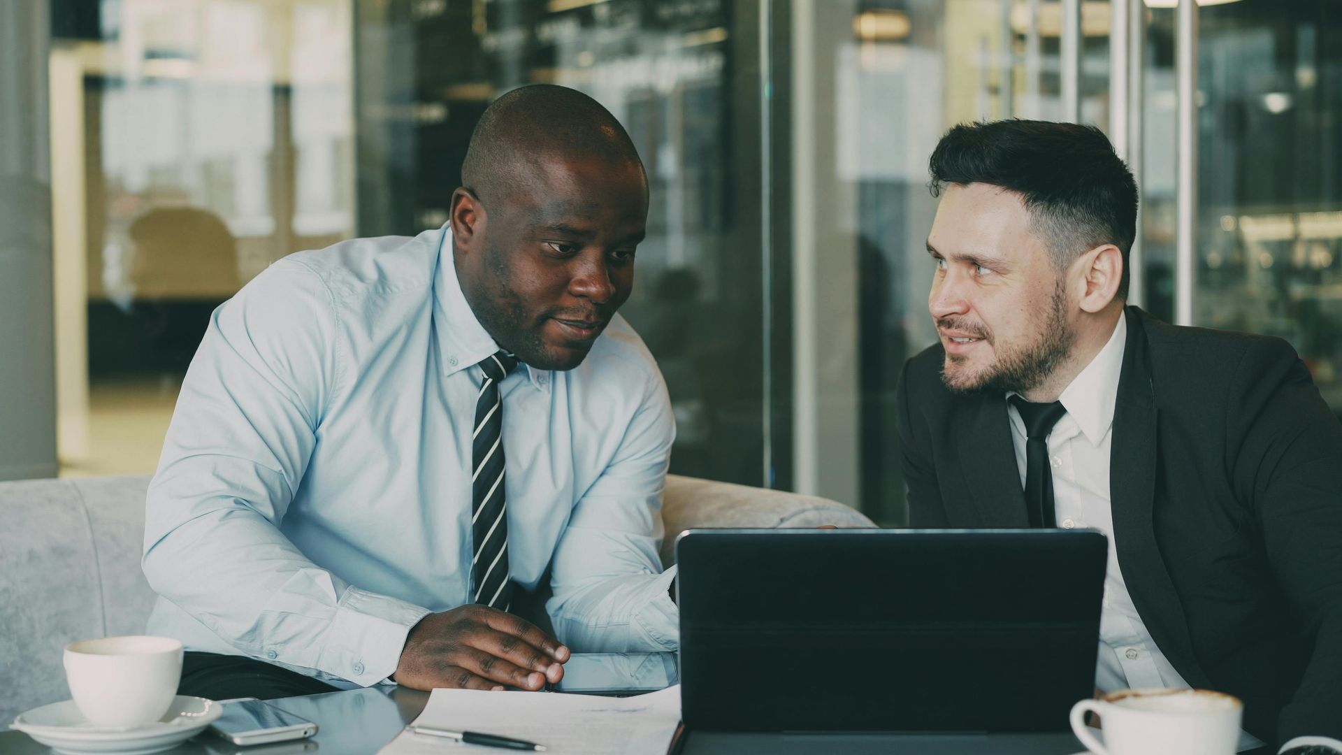 Two professionals in suits talking across a table with a laptop in a glass-walled office