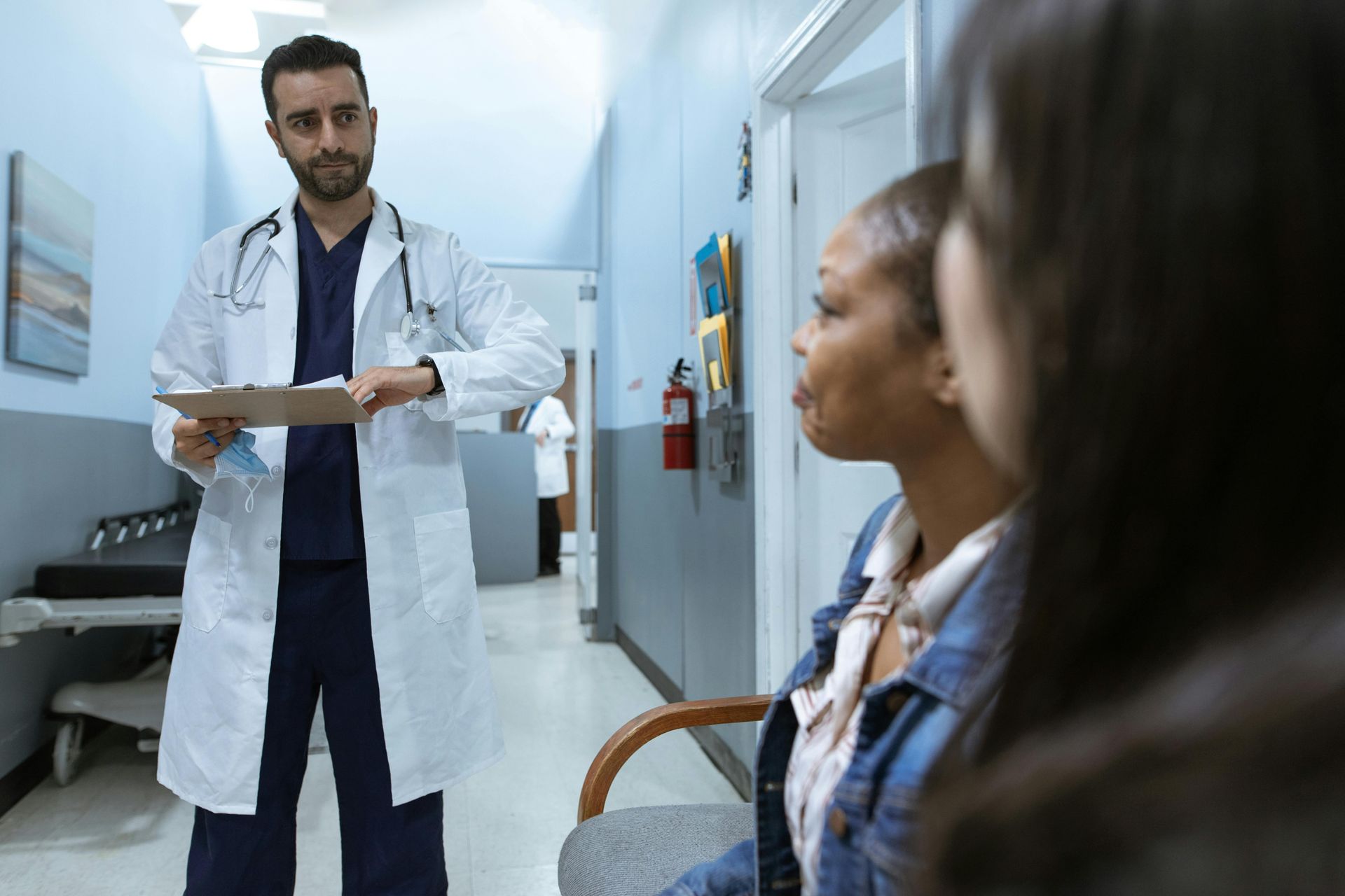Doctor speaking to two seated patients in a hospital hallway, holding a clipboard.
